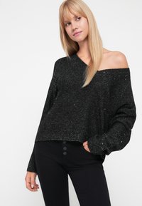 Maglione nero off-the-shoulder con una vestibilità ampia, texture scintillante e orlo a coste. Abbinato a pantaloni neri a vita alta con dettagli dei bottoni.