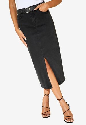 Kvinde iført en sort højtaljet denim midi-nederdel med frontslids, sort bælte, hvid top og sorte rem-sandaler, der krydser benene.