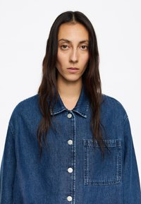Giacca di jeans blu scuro con colletto a camicia, chiusura anteriore con bottoni, una tasca sul petto e vestibilità rilassata. Texture liscia con leggera sbiaditura.