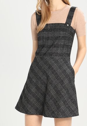 Kvinde iført en ærmeløs, sort og grå ternet romper over en kortærmet, ribbet beige top, stående med den ene hånd i lommen.