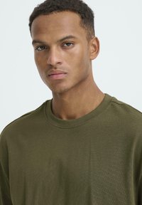 Tricou cu guler rotund de culoare verde măsliniu, fabricat dintr-un material moale și texturat, cu mâneci scurte. Prezintă o croială lejeră și detalii minime de cusătură.