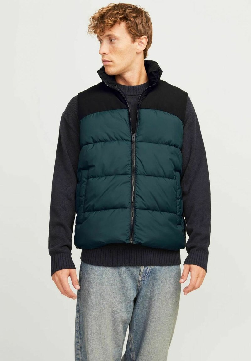 Gilet verde con pannello superiore nero, chiusura con zip e colletto alto. Indossato sopra un maglione scuro, abbinato a jeans chiari. Design casual.