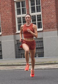 Løpeantrekk i rustrød: ermeløs croptopp og shorts. Atletiske sko i lyse røde med gule detaljer. Asfaltert gate og murstein bakgrunn.