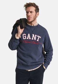 Navyblauwe sweatshirt met "GANT" in rode tekst. Heeft een ronde hals, geribbelde manchetten en zoom. G gedragen over een geruiten shirt, vastgehouden met een jas.