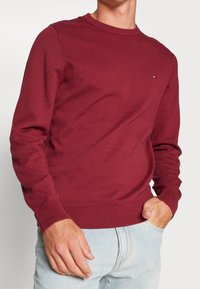 Maglione girocollo bordeaux, con polsini e fondo a costine, realizzato in tessuto morbido. Dettaglio del piccolo logo sul lato sinistro del petto, abbinato a jeans azzurri chiari.