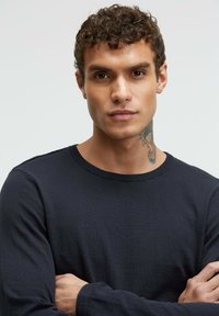 Marinefarbener Pullover mit langen Ärmeln und Rundhalsausschnitt aus Strickstoff; glatte Textur mit gerippten Bündchen; zeigt ein sichtbares Tattoo am Hals.