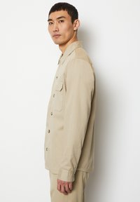 Beige button-up shirt with long sleeves, single chest pocket, and a flat collar; made from smooth fabric with a casual fit.