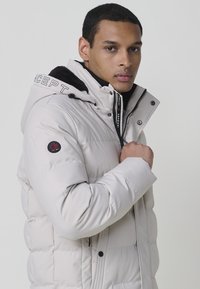 Ľahká puffrová bunda v off-white farbe, s odnímateľnou kapucňou s čiernym podšívkou, predným zipsom a vyšitým logom na rukáve.