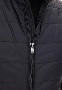 Närbild av en person som bär en mörk vadderad jacka med en silverfärgad dragkedja som är delvis stängd över en svart tröja.