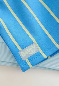 Blauw stof met lichtgele verticale strepen en een genaaide patch met het label "Bluey", geplaatst over lichtblauw geribbeld stof.