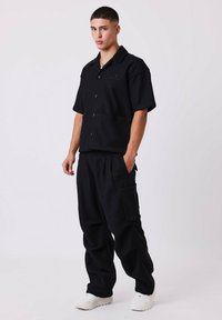 Camicetta nera a maniche corte con colletto e due bottoni, abbinata a pantaloni neri larghi con tasca laterale e sneakers bianchi.