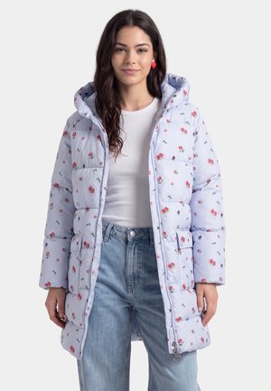 Junge Frau trägt eine hellblaue gesteppte Jacke mit floralem Muster, ein weißes geripptes Top, blaue Jeans und herzförmige rote Ohrringe, steht vor einem schlichten Hintergrund.