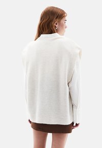 Pull sans manches beige clair avec un col et un ourlet côtelés. Présente une texture douce, porté sur une chemise blanche à manches longues.