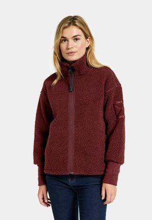Kvinde står og vender fremad iført en vinrød sherpa fleecejakke med lynlås og blå jeans mod en ensfarvet lys baggrund.