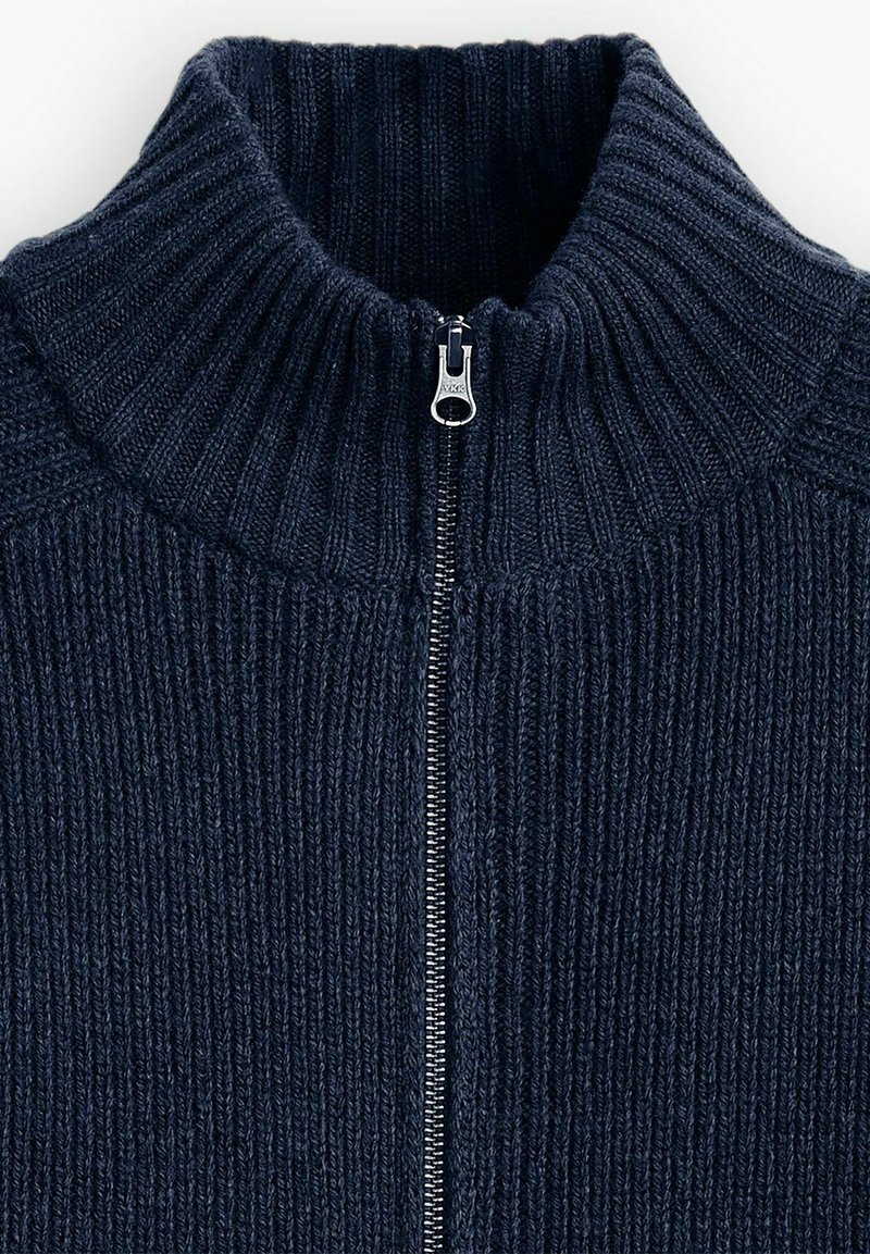 Suéter de punto azul marino con un cuello alto y cremallera plateada de longitud completa. Presenta una textura acanalada y líneas verticales a lo largo de la tela.