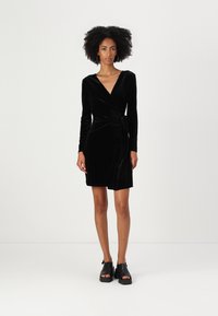 Vestido envolvente de veludo preto com mangas longas, decote em V, detalhe de cintura atada e comprimento curto, combinado com sandálias plataforma pretas.