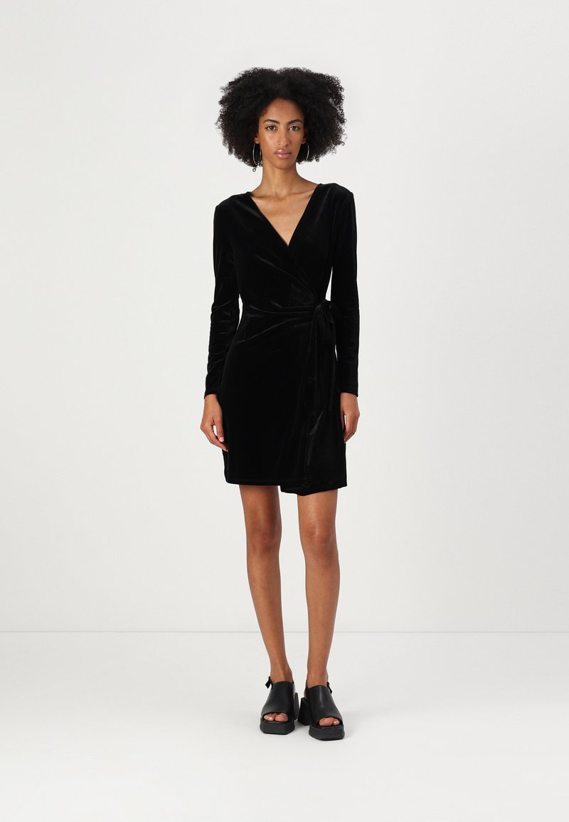 Vestido envolvente de veludo preto com mangas longas, decote em V, detalhe de cintura atada e comprimento curto, combinado com sandálias plataforma pretas.