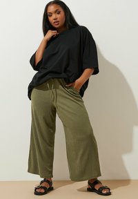 T-shirt noir surdimensionné associé à un pantalon large vert olive avec une finition texturée et un cordon de serrage. Sandales noires avec une boucle décorative.