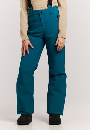 Persoon draagt teal snowpants met zwarte bretels, beige top met lange mouwen en zwarte winterlaarzen, staand tegen een effen achtergrond.