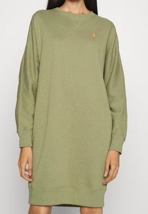 Frau trägt ein übergroßes, olivgrünes Sweatshirt-Kleid mit langen Ärmeln und einem kleinen orangefarbenen gestickten Logo auf der linken Brust.