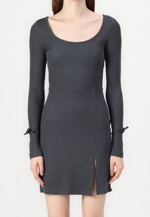 Kvinde iført en tætsiddende mørkegrå langærmet kjole med scoop-halsudskæring, snore ved håndleddene og et frontskridt i nederdelen.