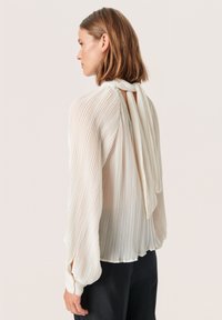 Transparante, geplooide blouse in off-white met een hoge hals en een strik aan de achterkant. Met lange mouwen en een losse pasvorm. Gestructureerde stof.