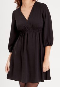 Robe noire avec un décolleté en V, des manches bouffantes et une taille élastique. Le tissu est lisse, avec une jupe évasée arrivant à mi-cuisse.