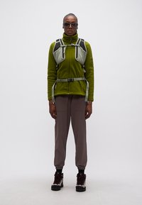 Άτομο που φοράει πράσινο φλις μπουφάν, γκρι σακίδιο με ιμάντες, καφέ παντελόνι cargo, μπότες πεζοπορίας και γυαλιά ηλίου, στέκεται μπροστά σε λευκό φόντο.