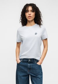 Ανοιχτό μπλε βαμβακερό μπλουζάκι με κοντά μανίκια, στρογγυλή λαιμόκοψη, με μικρό κεντημένο λογότυπο σε ναυτικό μπλε στο αριστερό στήθος. Συνδυασμένο με μπλε τζιν.