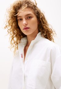 Camicia bianca con bottoni e tasca, dotata di colletto piegato e ricamo rosso "S.R.G." sulla pattina. Tessuto di cotone morbido.