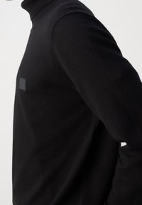 Pull à col roulé noir avec un col côtelé, confectionné en tissu tricoté doux. Il présente un discret écusson logo sur la poitrine.