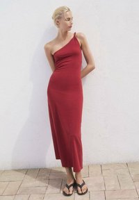 Rochie midi asimetrică, de culoare roșie, confecționată dintr-un material elastic, cu un singur bretele pe umăr și o siluetă ajustată. Purificată cu sandale negre.