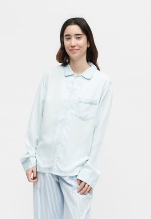 Γυναίκα με σκούρα μαλλιά φοράει ανοιχτό μπλε πιτζάμα με κουμπιά και ριγέ παντελόνι πιτζάμα, στέκεται μπροστά σε απλό φόντο.