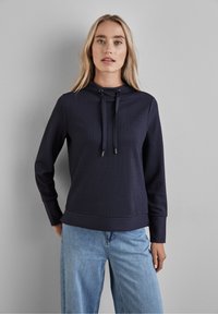 Granatowa, prążkowana bluza z wysokim dekoltem, ze sznurkiem i długimi rękawami, w zestawie z jasnoniebieskimi dżinsami.