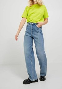 Person med en neongrön kortärmad skjorta, högmidjade ljusblå raka jeans och svarta klumpiga skor, står på ett ljust golv.