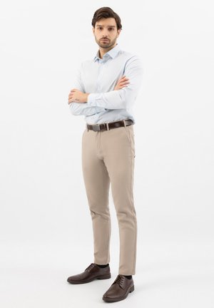 Man met kort donker haar die een lichtblauw overhemd met knopen draagt, beige broek, bruine riem en bruine nette schoenen, staand met gekruiste armen.