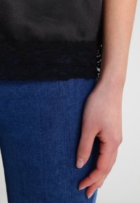 Haut en satin noir avec bordure en dentelle à l'ourlet, associé à un jean denim bleu foncé. Une main reposant sur le jean montre un teint de peau naturel.