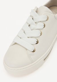 Tenis alb din piele, cu vârf rotund, șireturi albe și ochiuri aurii. Prezintă o talpă din cauciuc texturat și detalii minime de cositură.