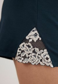Mörk teal-plagg med en dekorativ spetspanel med blommönster och ett nätbakgrund vid fållen. Texturerad yta nära kanten.