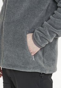 Giacca in fleece grigia con tasca con zip, tessuto strutturato e bordi a contrasto ai polsi, abbinata a pantaloni neri.