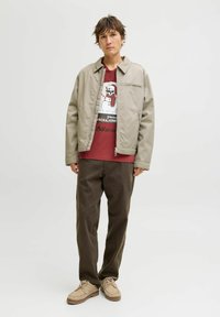 Lichtbeige jas met ritssluiting, kastanjebruine t-shirt met een sneeuwpop afbeelding, bruine broek en tan kleurige schoenen met veters.