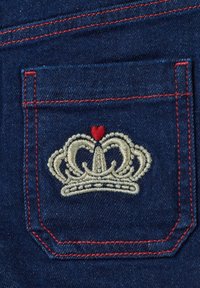 Bolsa de mezclilla con bordado de una corona dorada, que incluye un pequeño corazón rojo. Acentos de costura roja sobre un fondo de tela azul oscuro.