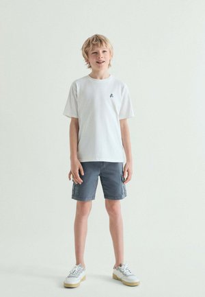 Niño rubio con camiseta blanca, pantalones cortos cargo grises y zapatillas blancas, de pie frente a un fondo claro y liso, mirando ligeramente hacia arriba y sonriendo.