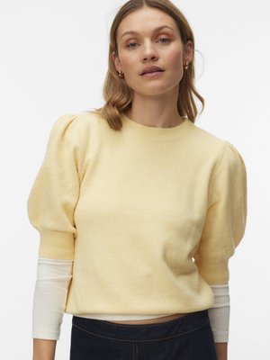 Femme portant un pull jaune clair à manches courtes avec des épaules bouffantes, par-dessus un haut blanc à manches longues et un pantalon foncé, sur un fond uni.