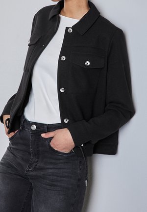 Persoon draagt een zwart jasje met knopen over een wit topje en donker gewassen jeans, handen in de zakken, staande tegen een lichtgrijze muur.