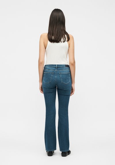 Γυναίκα που φοράει λευκό αμάνικο τοπ και μπλε τζιν παντελόνι bootcut, στέκεται με την πλάτη στραμμένη προς τα εμπρός σε απλό φωτεινό φόντο.