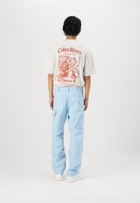 Pantalones cargo de color azul claro con múltiples bolsillos, combinados con una camiseta beige que presenta un diseño gráfico rojo. Corte simple y relajado.