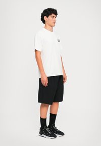 Jovem homem de pé, vestindo uma t-shirt branca, calções pretos até ao joelho, socks pretas e ténis preto e branco, em frente a um fundo liso.