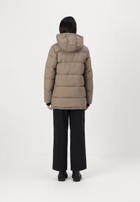 Personne portant une doudoune taupe à capuche, un pantalon noir à jambes larges et des chaussures plateformes noires, debout de dos sur un fond blanc.