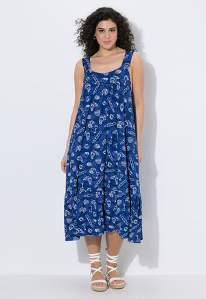 Kvinde med krøllet hår iført en ærmeløs blå midi-kjole med hvide bager-tema prints og hvide snøresandaler.